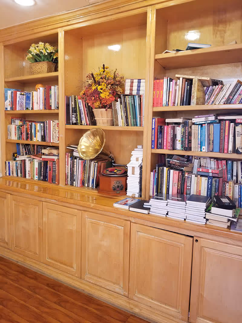 Wooden built-in bookshelf filled with various books, a vintage gramophone, a flower arrangement in a basket, and stacks of notebooks, set against a polished wooden floor.