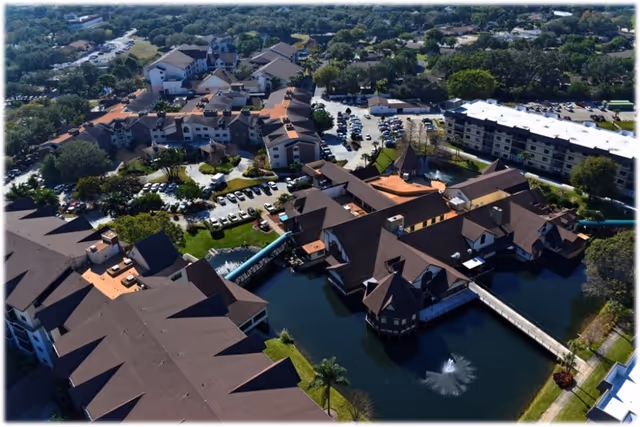 Aerial view of The Inn at Freedom Village senior living facility showing multiple buildings with brown roofs surrounding a pond with a fountain, parking lots with cars, and green landscaped areas with trees.
