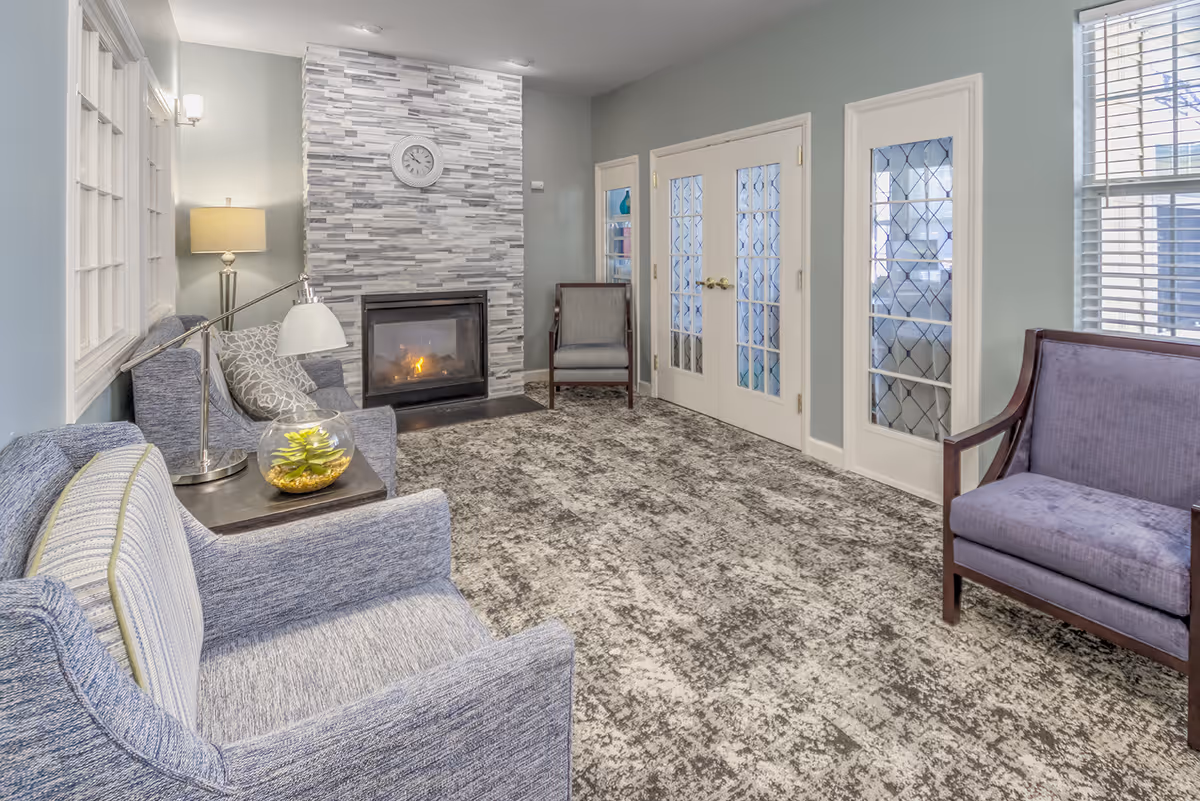 A cozy living room with a modern fireplace surrounded by gray stone tiles. The room features several upholstered chairs in shades of blue and gray, a side table with a lamp and a small plant, a patterned carpet, and French doors with decorative glass panels. The walls are painted light blue, and there are windows with white blinds allowing natural light into the room.