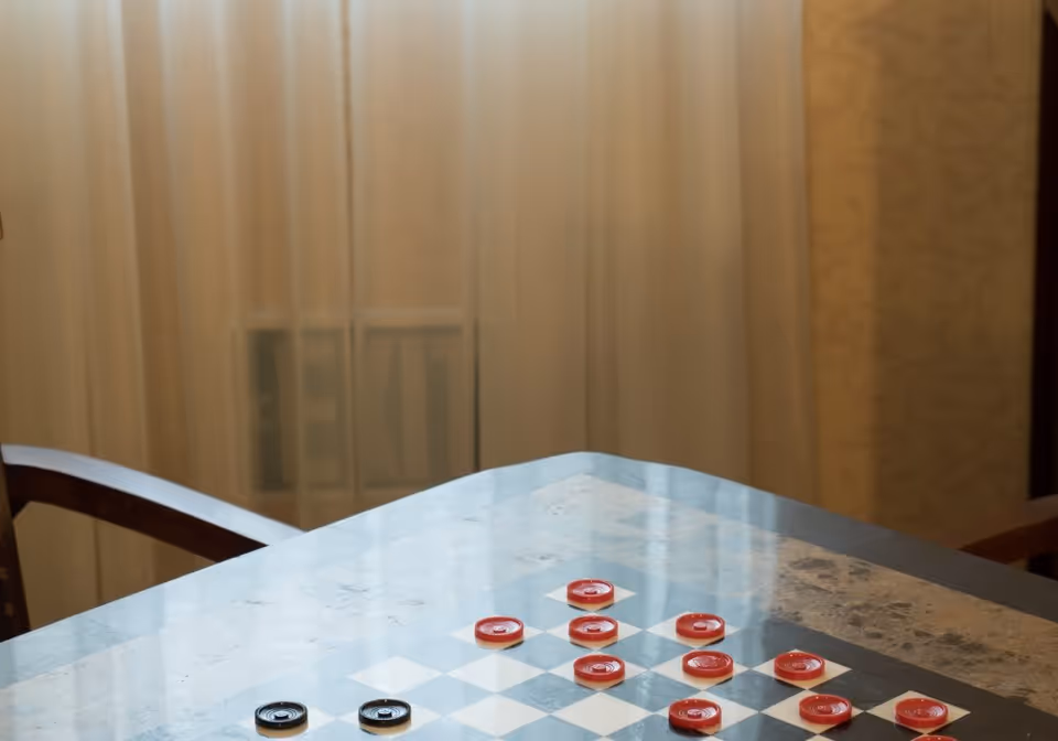 A checkers game set up on a glass-topped table with red and black checkers pieces. The table is indoors with a sheer curtain and a wooden chair visible in the background.