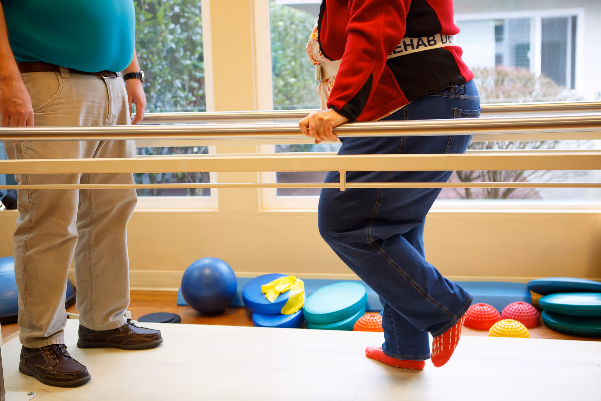A person wearing a red jacket and blue jeans is holding onto parallel bars while performing a balance exercise on one foot. Another person stands nearby, wearing beige pants and brown shoes. Various exercise equipment, including balance pads and balls, are visible in the background near a window.