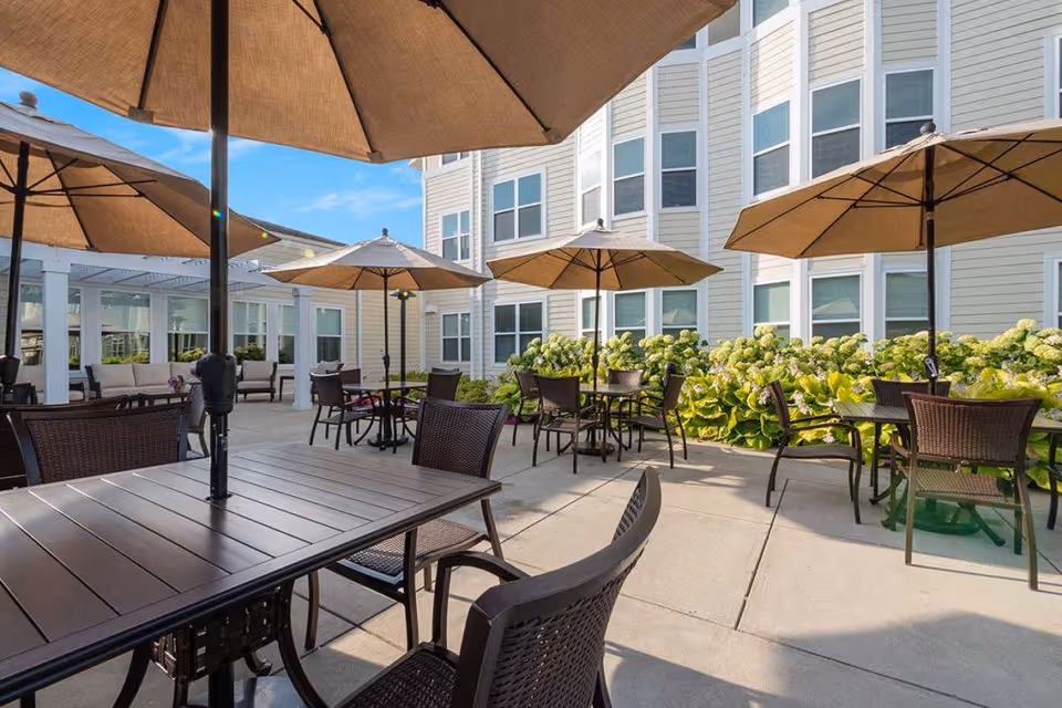 Outdoor patio area at EverTrue Concordia Village with multiple tables and chairs under large beige umbrellas, surrounded by green plants and the exterior of a multi-story building with many windows.