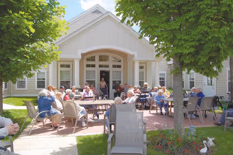 A group of elderly people sitting and socializing on patio chairs and benches outside a light-colored assisted living residence building. The area is shaded by trees and has a brick-paved ground with some flower beds nearby.