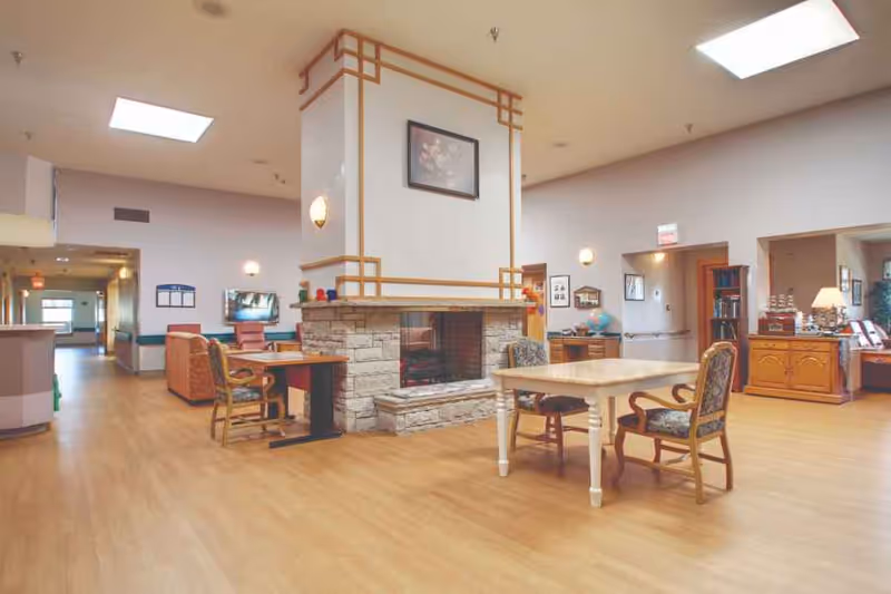 A spacious common area in a senior living facility featuring a central stone fireplace with a framed floral picture above it. The room has wooden flooring, several tables with chairs, a bookshelf, and soft lighting from wall sconces and ceiling lights. The walls are painted in light colors, and there are doorways leading to other rooms.