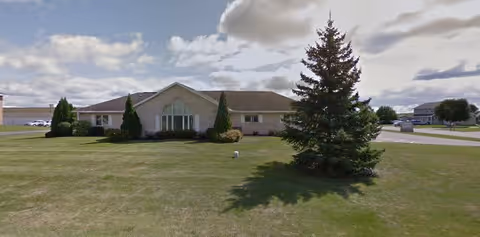 A single-story building with a light-colored exterior and a gabled roof, surrounded by a well-maintained lawn with a large evergreen tree and some shrubs. The sky is partly cloudy.