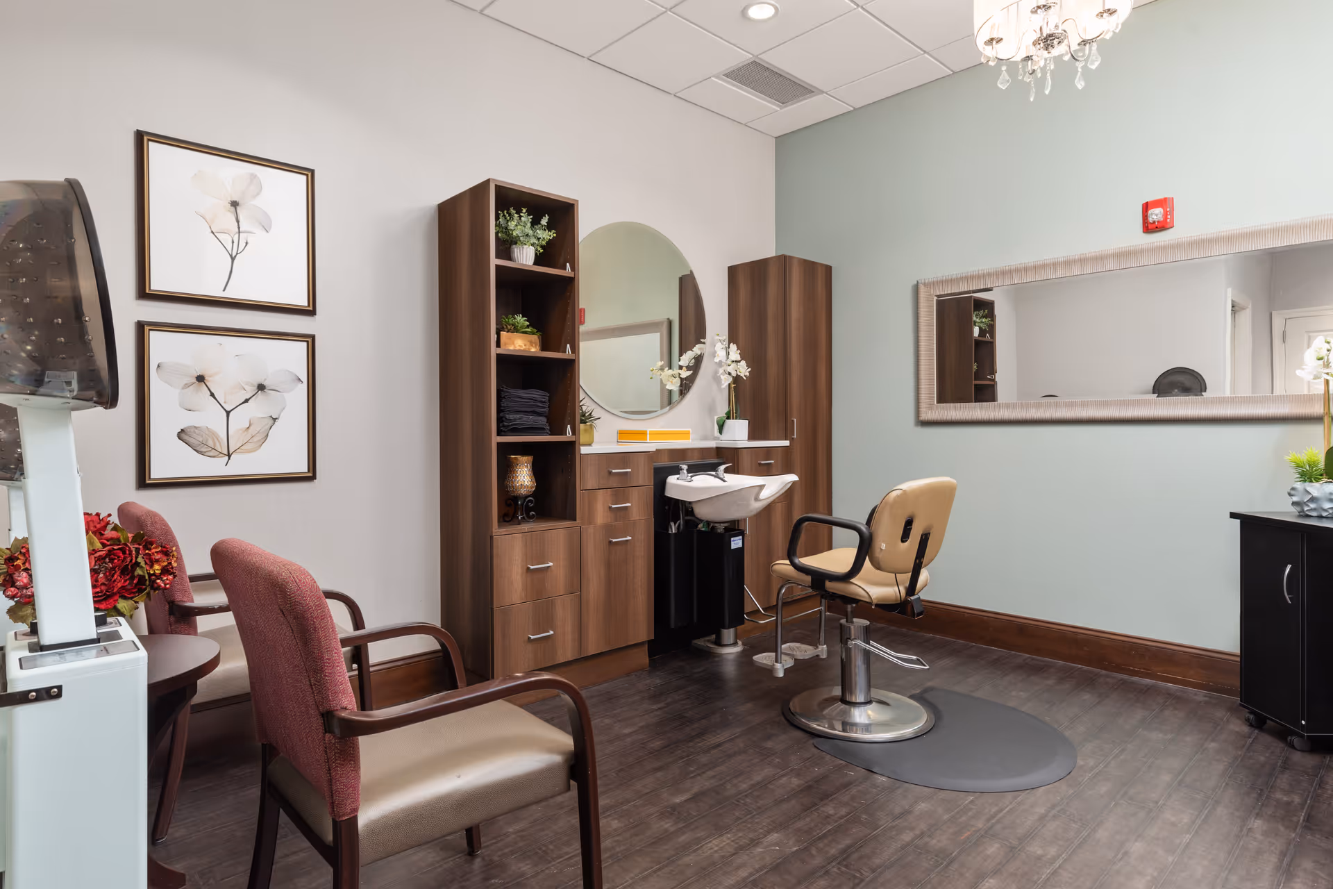 A well-lit salon area in an assisted living facility featuring a salon chair in front of a sink and vanity with a large oval mirror. There are wooden cabinets and shelves with decorative plants and towels. Two upholstered chairs are placed nearby, and a large rectangular mirror hangs on a light green wall. The floor is dark wood, and a chandelier light fixture hangs from the ceiling.