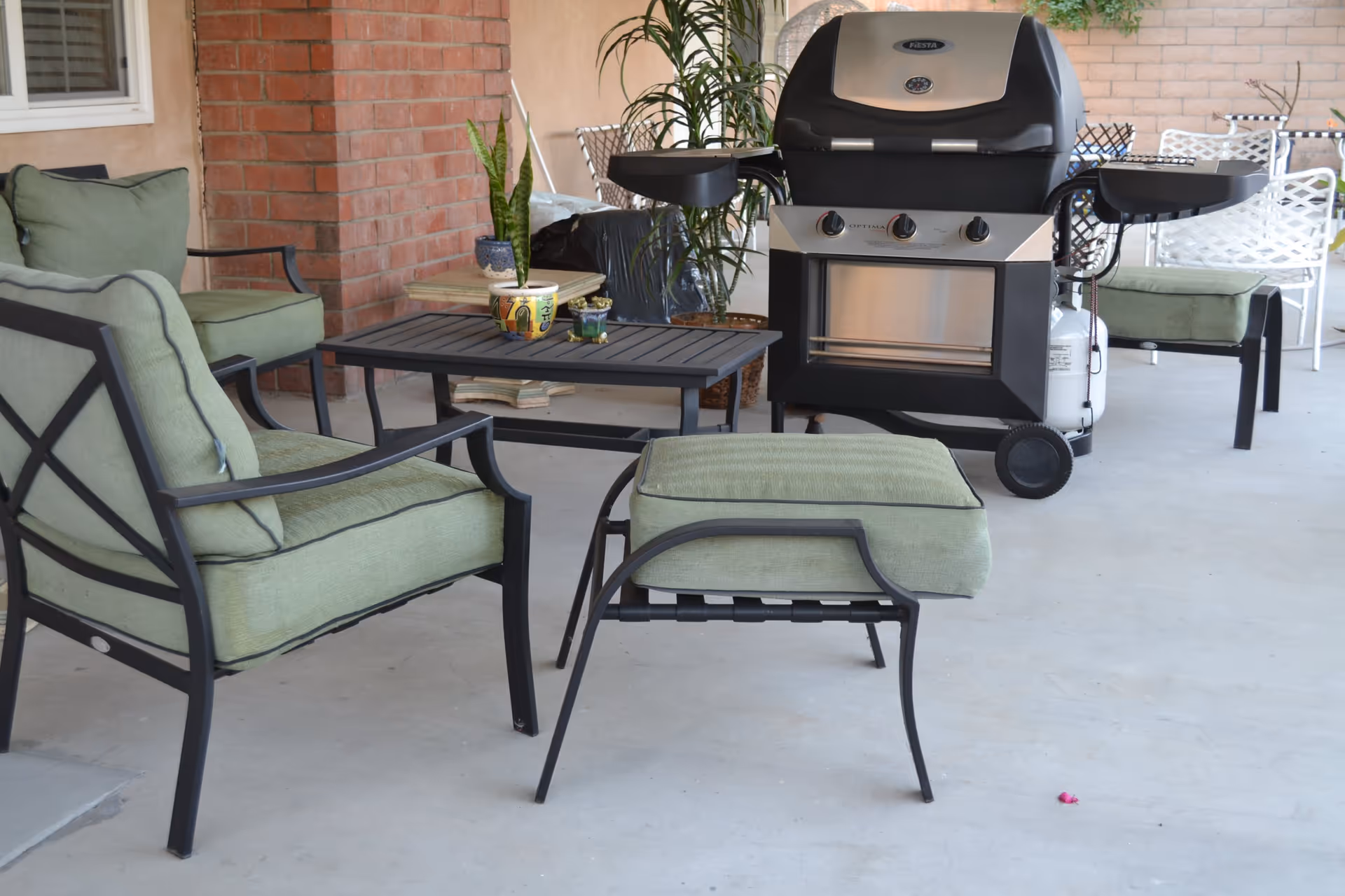 Outdoor patio area with green cushioned metal chairs and a matching ottoman, a black metal coffee table with potted plants, and a black and silver gas grill. There is a brick wall and a window in the background along with additional white metal chairs and a table.