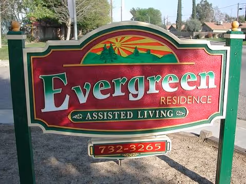 A decorative outdoor sign for Evergreen Residence Assisted Living, featuring a green and red color scheme with a sun and mountain graphic at the top, and a phone number 732-3265 displayed below.