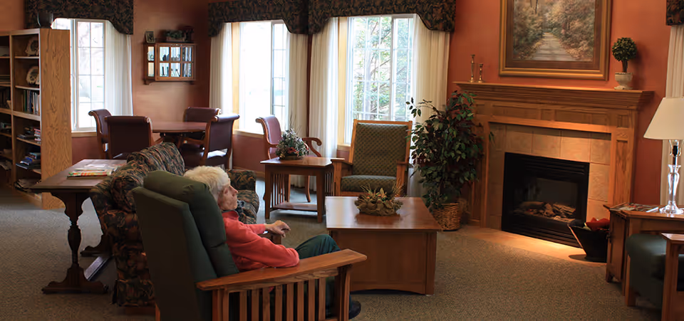 An elderly woman sits in a comfortable communal living room with armchairs, a coffee table, large windows, and a lit fireplace.