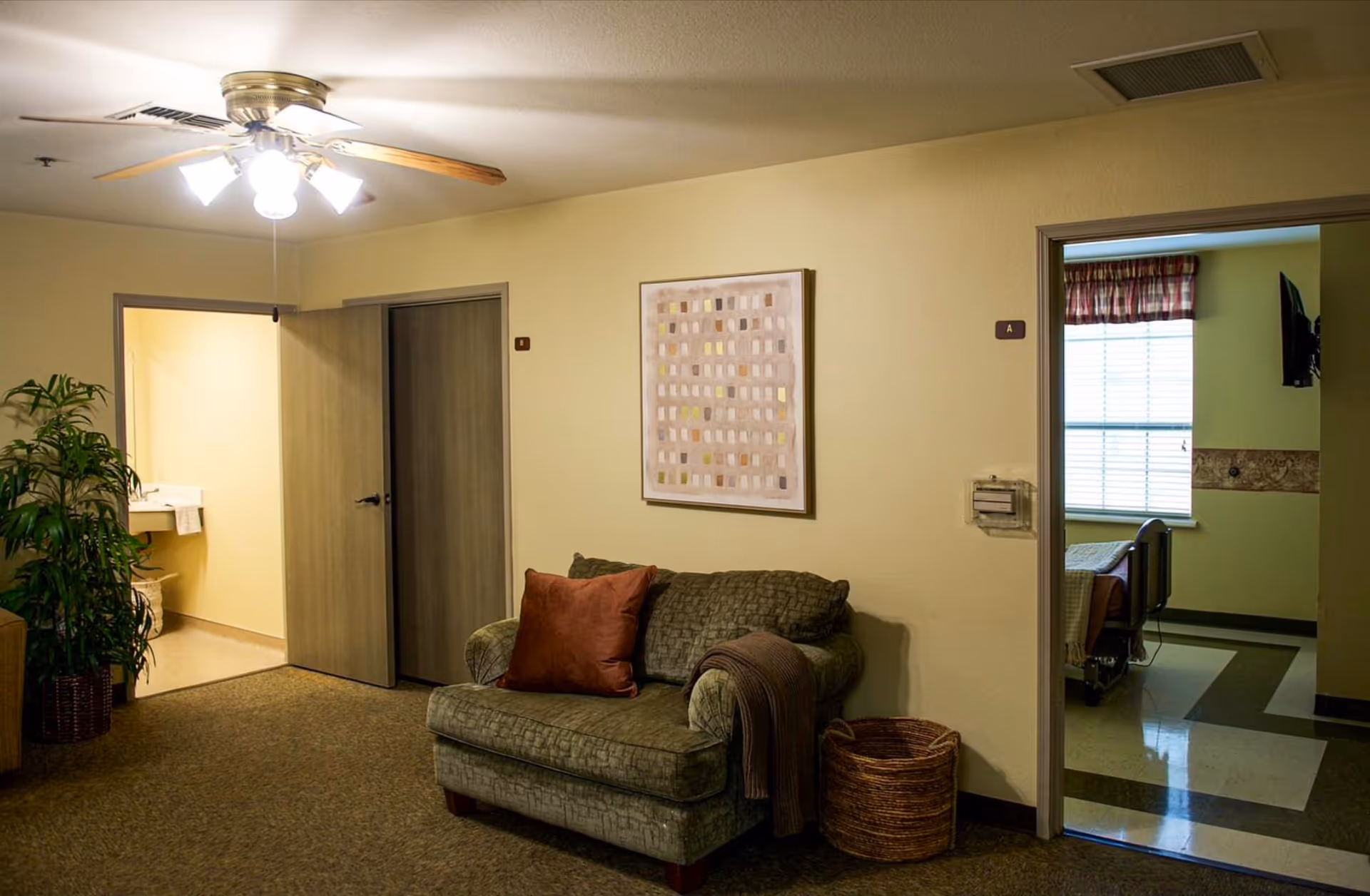 Small living area in a senior facility with a loveseat, ceiling fan, wall art, potted plant, and doorways leading to a bathroom and a bedroom with a hospital bed.