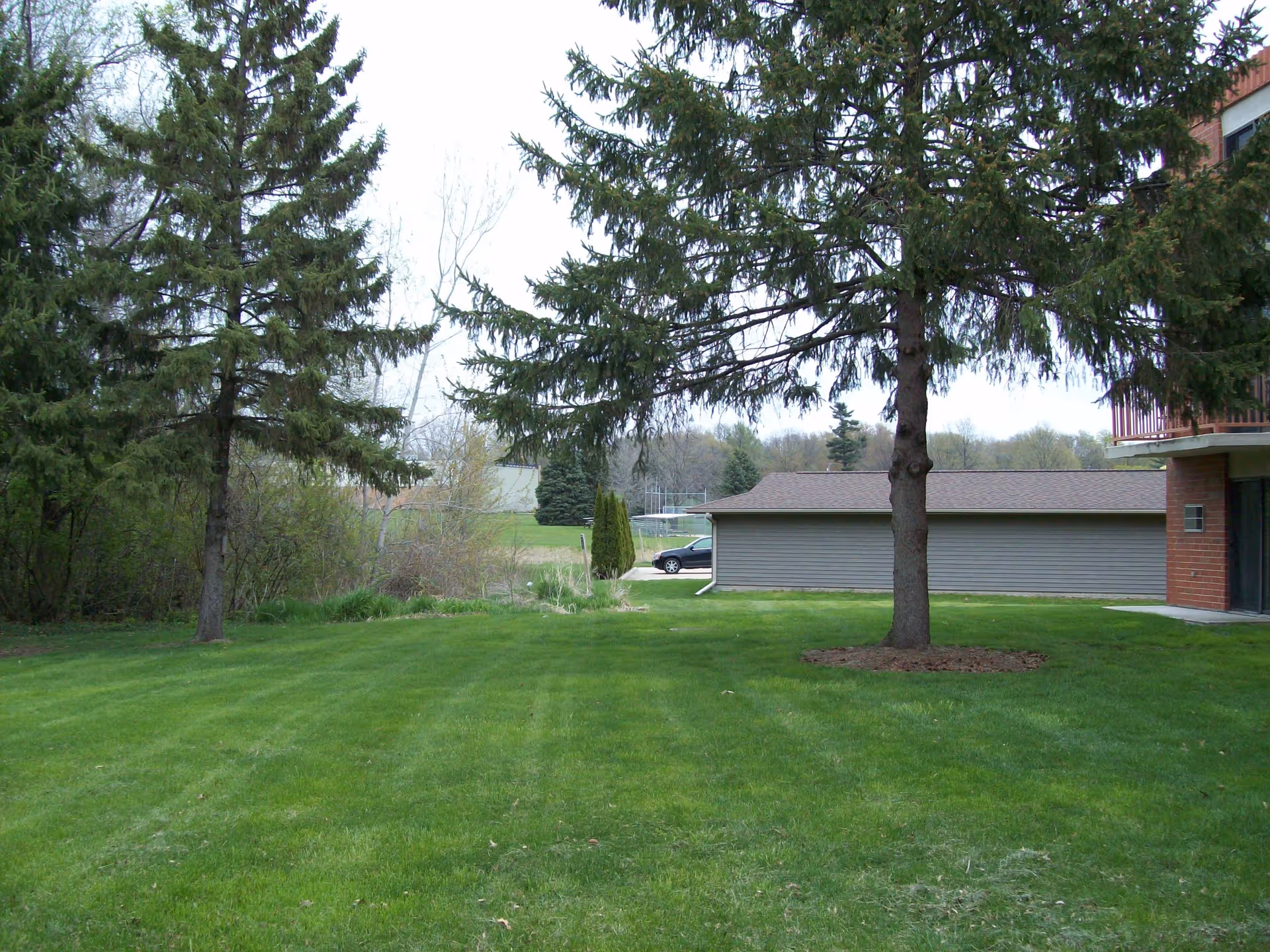 A grassy outdoor area with several tall evergreen trees, a small building with a sloped roof, and part of a brick building with a balcony on the right side.