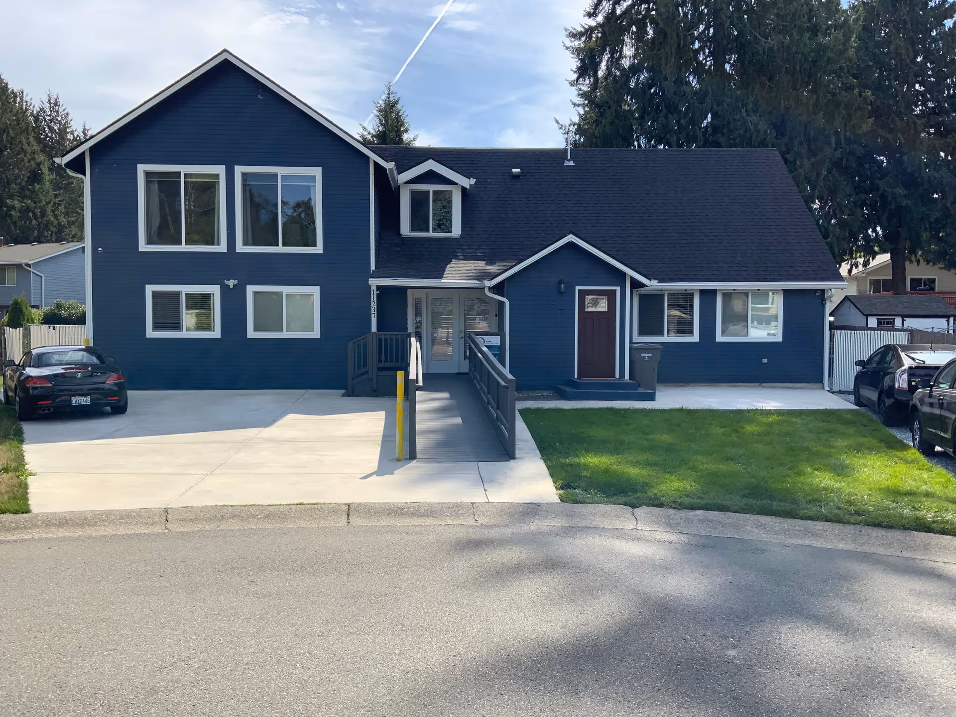 Front exterior view of a two-story dark blue house with white trim, featuring a wheelchair accessible ramp leading to the main entrance. There are several windows, a small porch with a brown door, a green lawn on the right side, and a concrete driveway with parked cars on both sides.