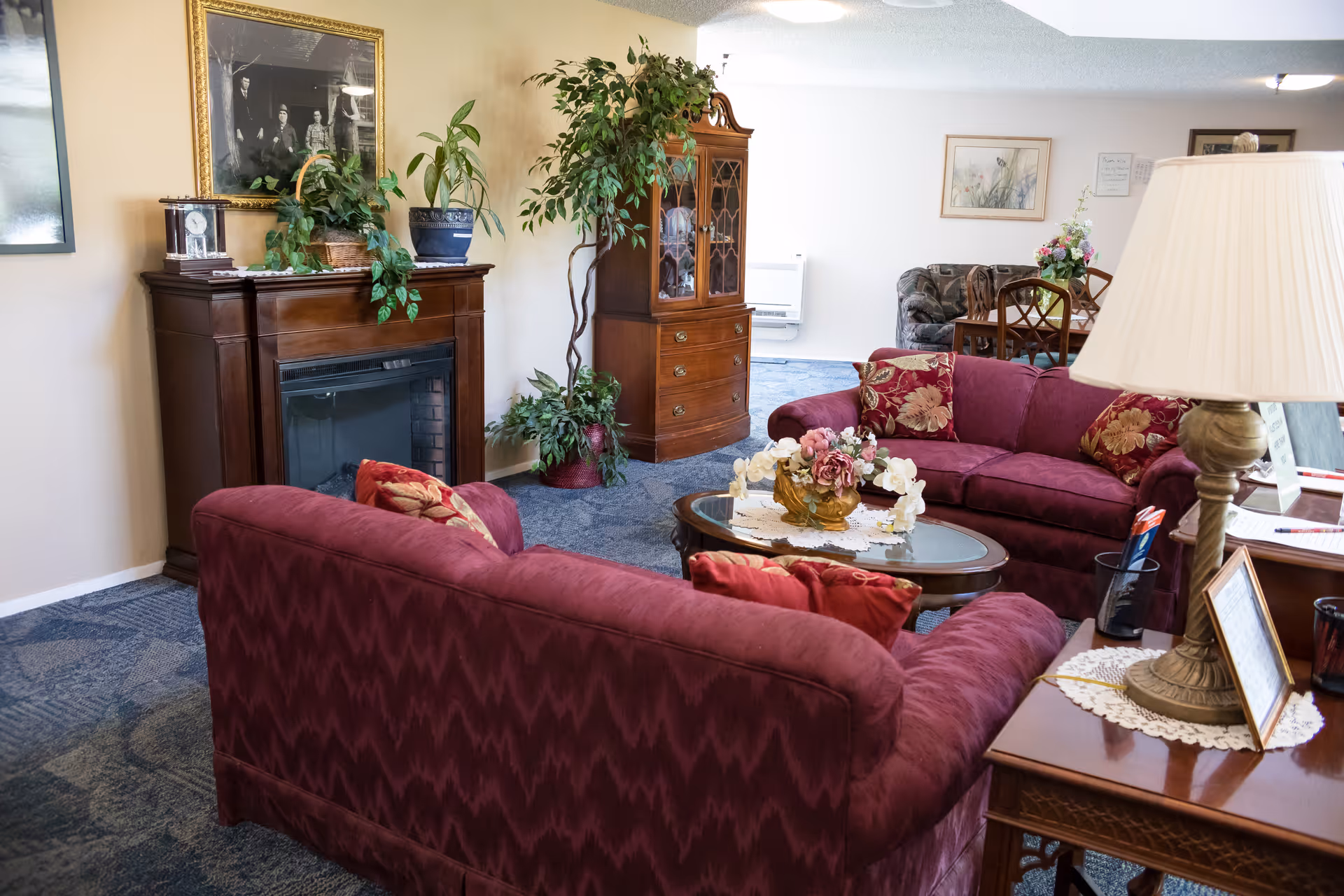 A cozy living room area with two maroon sofas adorned with floral cushions, a wooden coffee table with a floral centerpiece, a wooden side table with a lamp and framed photo, a wooden fireplace mantel with plants and a framed picture above it, and a wooden cabinet with glass doors. The room has blue carpet and light-colored walls.