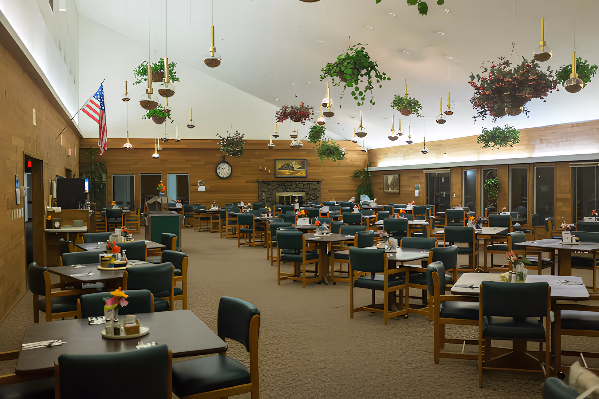 A spacious dining room with multiple tables and green cushioned chairs arranged neatly. The room features hanging plants and pendant lights from a high white ceiling. There is a stone fireplace on the far wall, an American flag on the left, and wood-paneled walls with large windows on the right side.