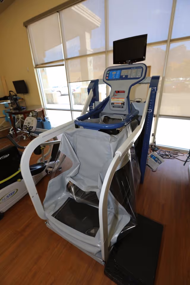A rehabilitation exercise machine with a seat enclosed in a transparent plastic cover, positioned in a room with large windows covered by beige roller shades. The machine has a digital control panel and a monitor on top, and the room has wooden flooring with other exercise equipment visible in the background.