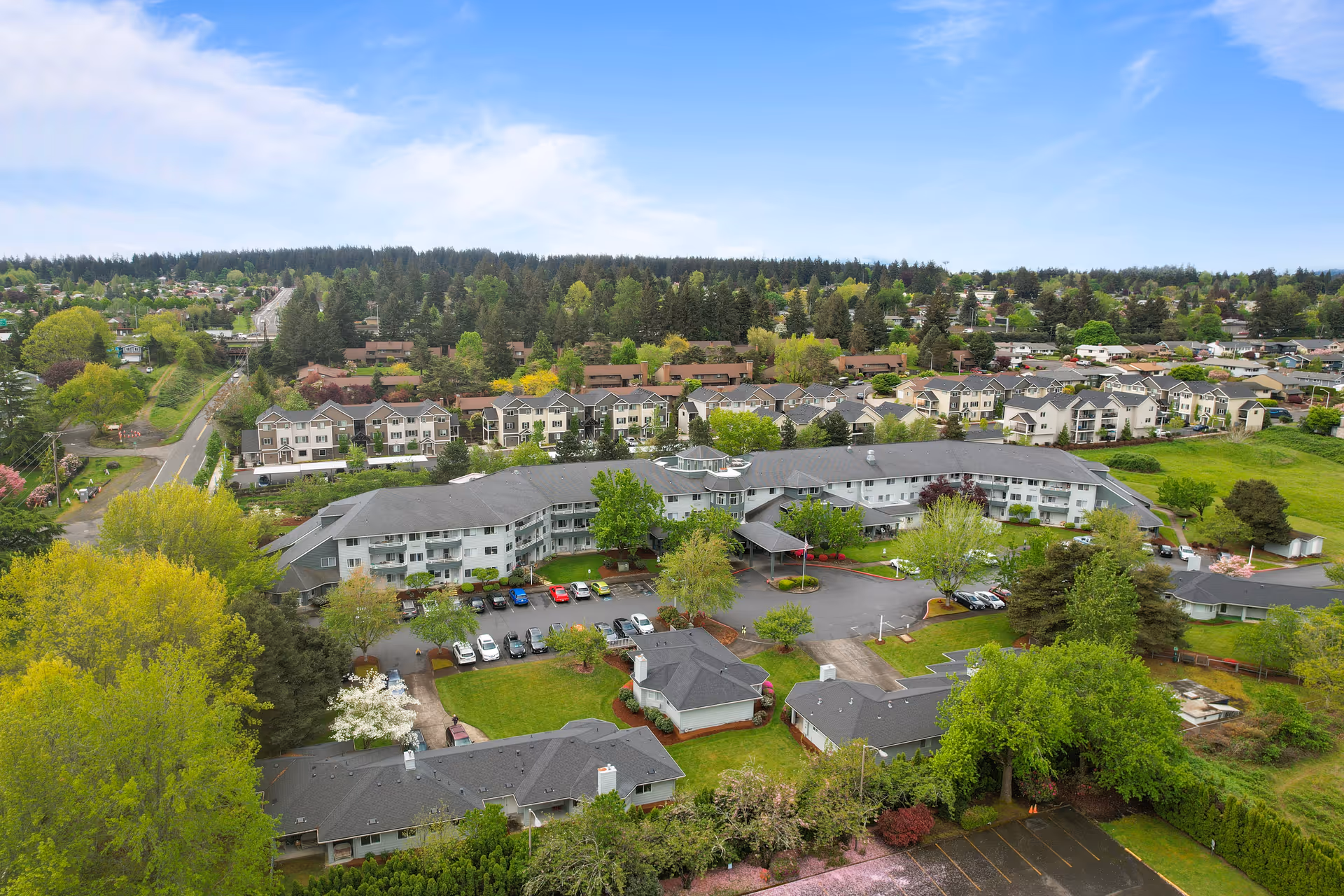 Aerial view of Parkrose Chateau Gracious Retirement Living facility surrounded by trees and greenery, with multiple buildings, parking lots, and a residential neighborhood in the background under a partly cloudy sky.