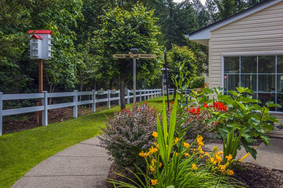 A landscaped outdoor walkway beside a light-colored building, bordered by colorful flowering plants, a white fence, a birdhouse, and trees.
