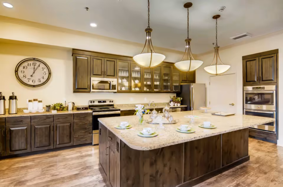 A spacious kitchen with wooden cabinets and a large central island topped with a granite countertop. The island is set with cups, saucers, and a silver tea set. Three pendant lights hang above the island. The kitchen features stainless steel appliances including a stove, microwave, refrigerator, and built-in oven. A large wall clock is mounted on the left wall.
