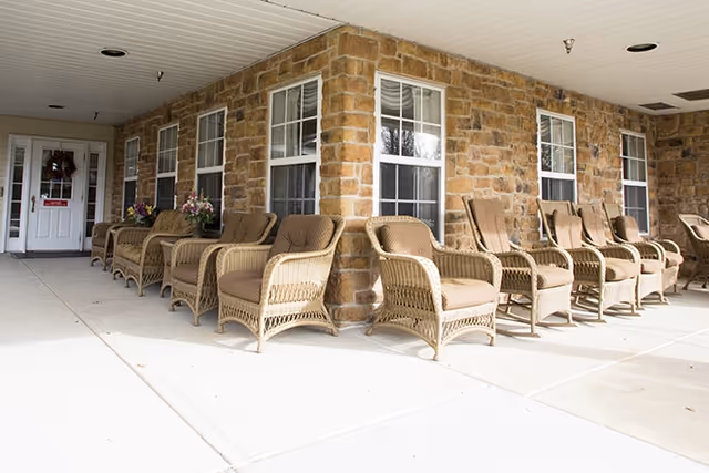 Covered front porch with a row of wicker chairs lined up against a stone exterior wall and windows.