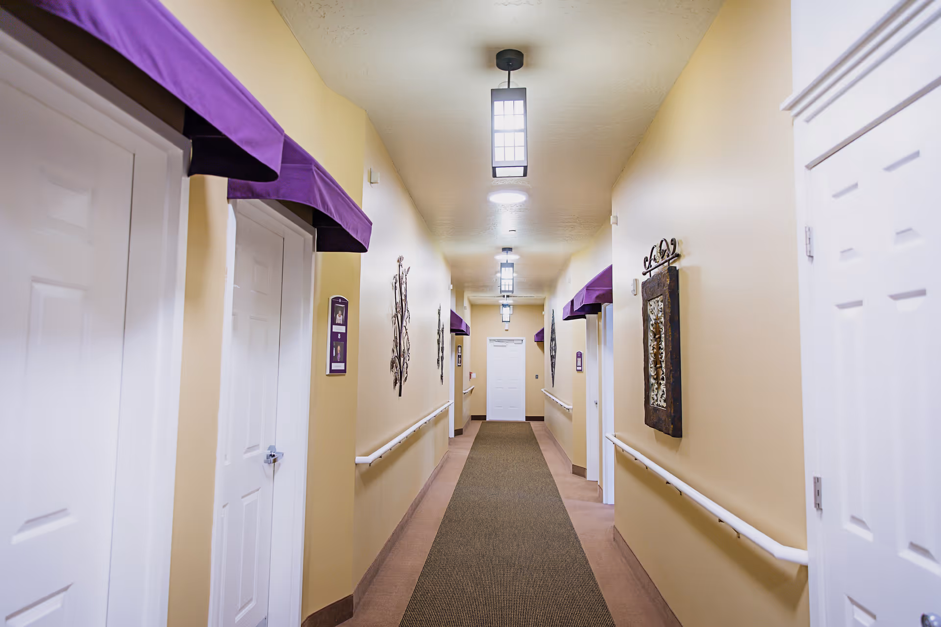 A well-lit indoor hallway with beige walls and a brown carpet runner. The hallway features white doors on both sides, each with a purple awning above. Handrails run along both walls, and decorative wall art is displayed intermittently. Ceiling lights are evenly spaced along the corridor.