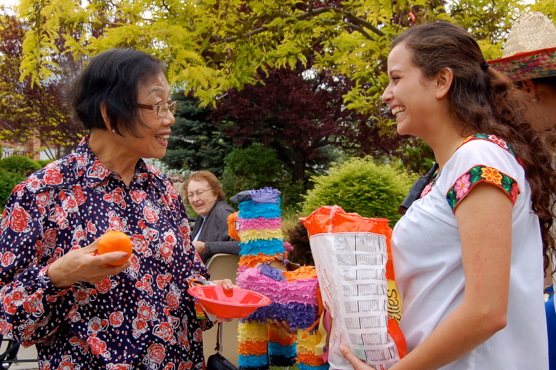 An elderly woman holding an orange and a red bowl smiling and talking to a younger woman who is holding a large bag of chips. They are outdoors with colorful trees and bushes in the background, and a small colorful piñata is visible between them. Another elderly woman is seated in the background, also smiling.