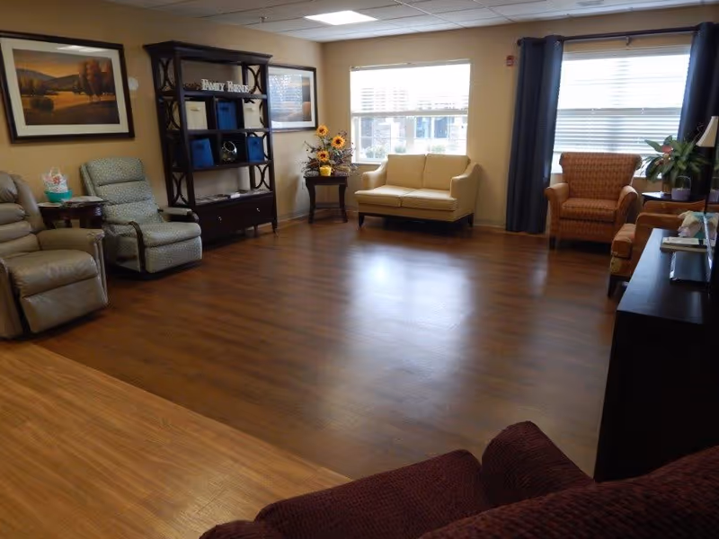A bright communal living room with sofas, armchairs, shelving, and large windows.