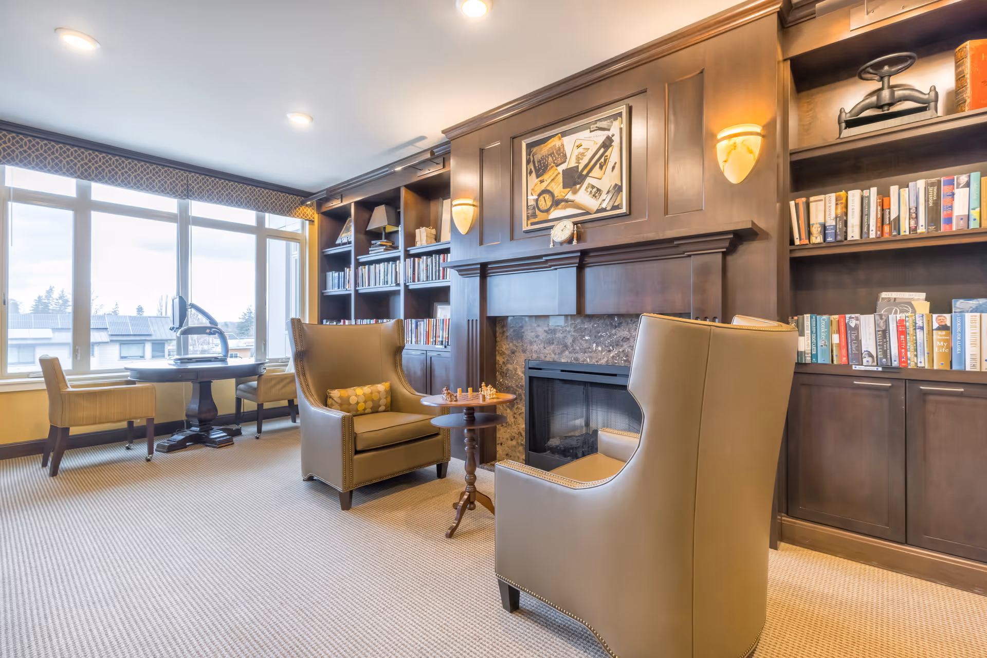 A cozy living room area with two beige armchairs facing a small round table with a chess set. Behind the chairs is a dark wood fireplace with built-in bookshelves filled with books and decorative items. Large windows on the left side let in natural light, and a round table with two chairs is positioned near the windows.
