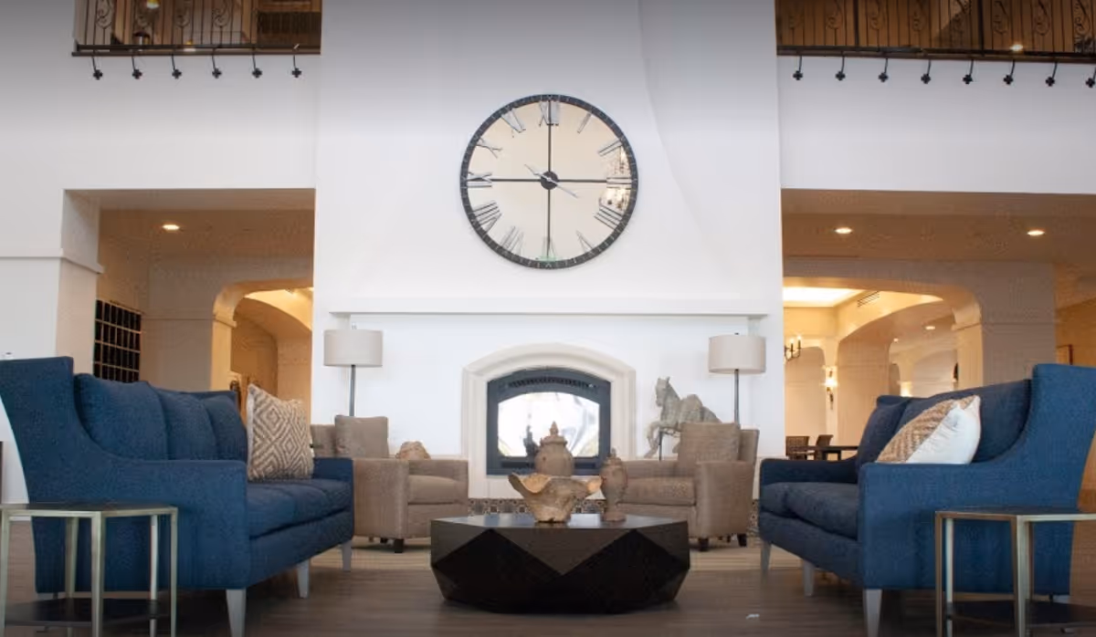 Spacious seating area with blue sofas and armchairs arranged around a central coffee table in front of a large wall clock and fireplace.