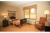 A well-lit bedroom in a senior living facility featuring a single hospital-style bed with a brown headboard and footboard, a bedside table with a lamp and phone, a comfortable armchair, a floor lamp, a window with a beige valance, and light-colored walls and flooring.
