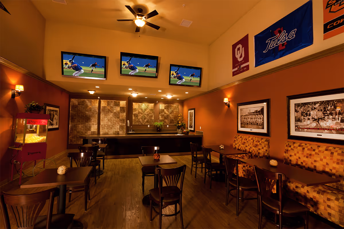 A cozy common area with multiple tables and chairs, a popcorn machine on the left, three wall-mounted TVs showing a baseball game, and sports banners and framed photos decorating the walls. The room has warm lighting and a wooden floor.