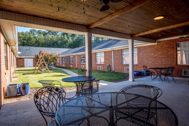 Covered outdoor courtyard with metal patio tables and chairs overlooking a grassy area and a brick building with a wooden swing.