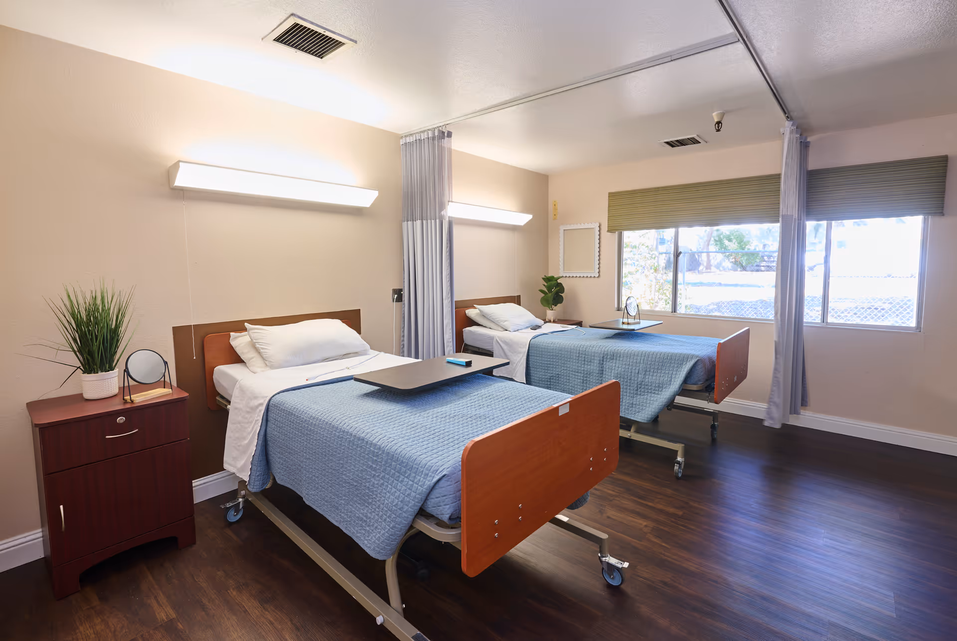 Two adjacent hospital-style beds with blue covers in a bright, well-lit senior care room with a nightstand and large windows.