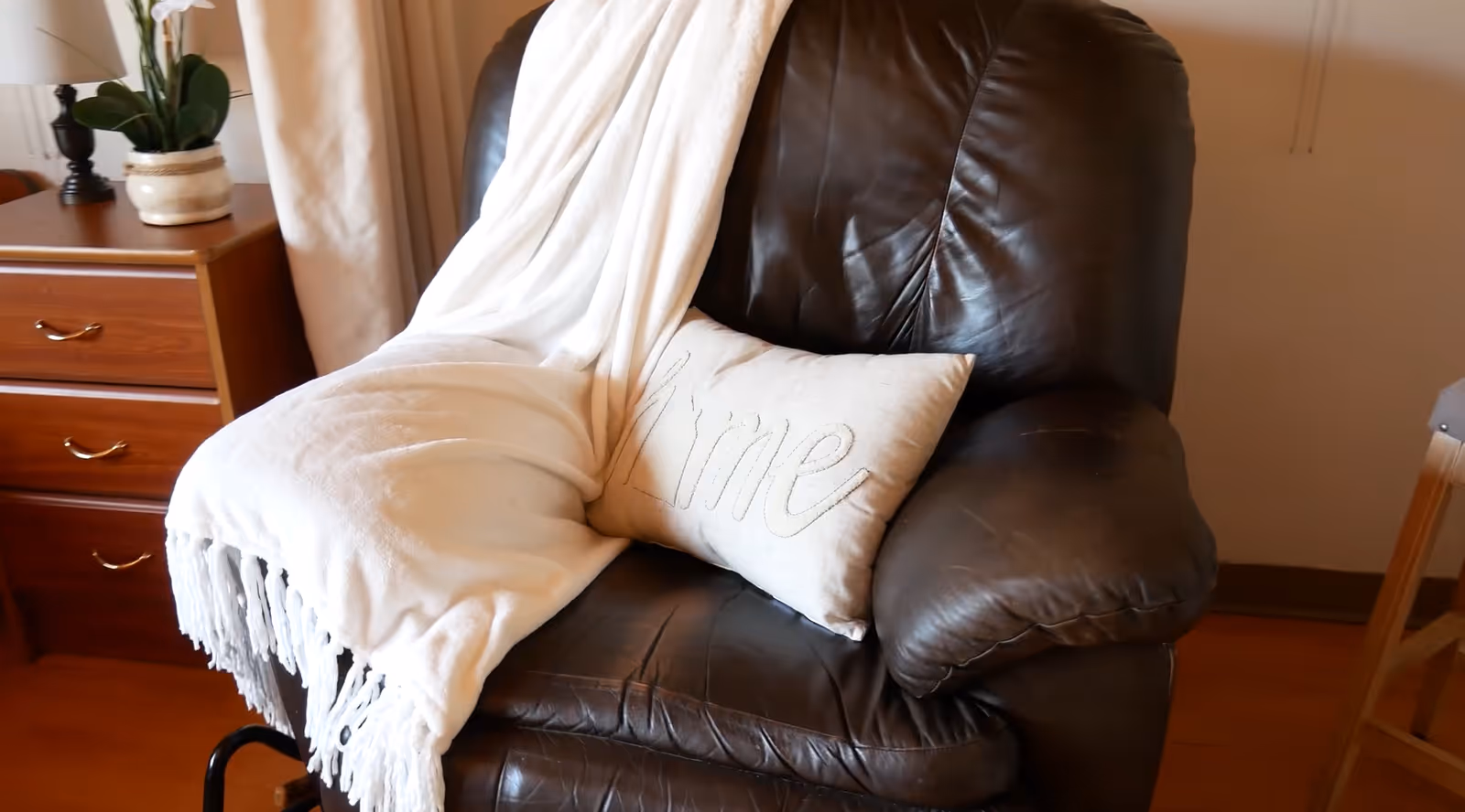A dark brown leather armchair with a white throw blanket draped over one side and a beige pillow with the word 'Home' embroidered on it. Next to the chair is a wooden nightstand with two drawers, a white lamp, and a potted plant with white flowers.