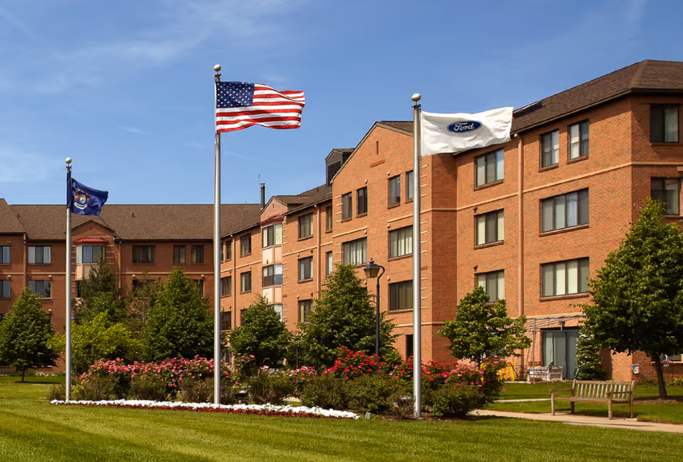 Exterior view of a multi-story brick building with several windows, surrounded by green grass, trees, and flower beds. Three flagpoles are visible in front of the building, flying the American flag, a state flag, and a Ford flag. A wooden bench is placed near a sidewalk on the right side.