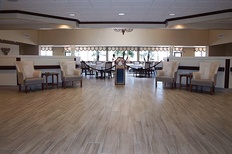 Open dining room in a senior living facility with dining tables and chairs seen beyond a tiled foyer lined with armchairs.