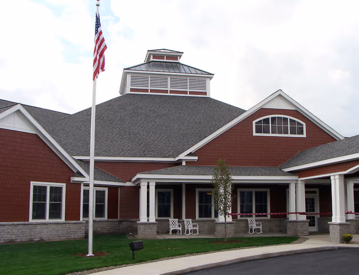 Exterior view of a red building with white trim, a gray shingled roof, and a covered porch area with white chairs. An American flag on a flagpole is visible in front of the building, along with a small tree and a curved driveway.
