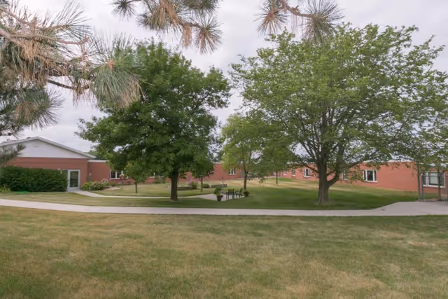 Outdoor view of Colonial Manor Of Randolph showing a grassy area with several trees, a paved walkway, and red brick buildings in the background under a cloudy sky.