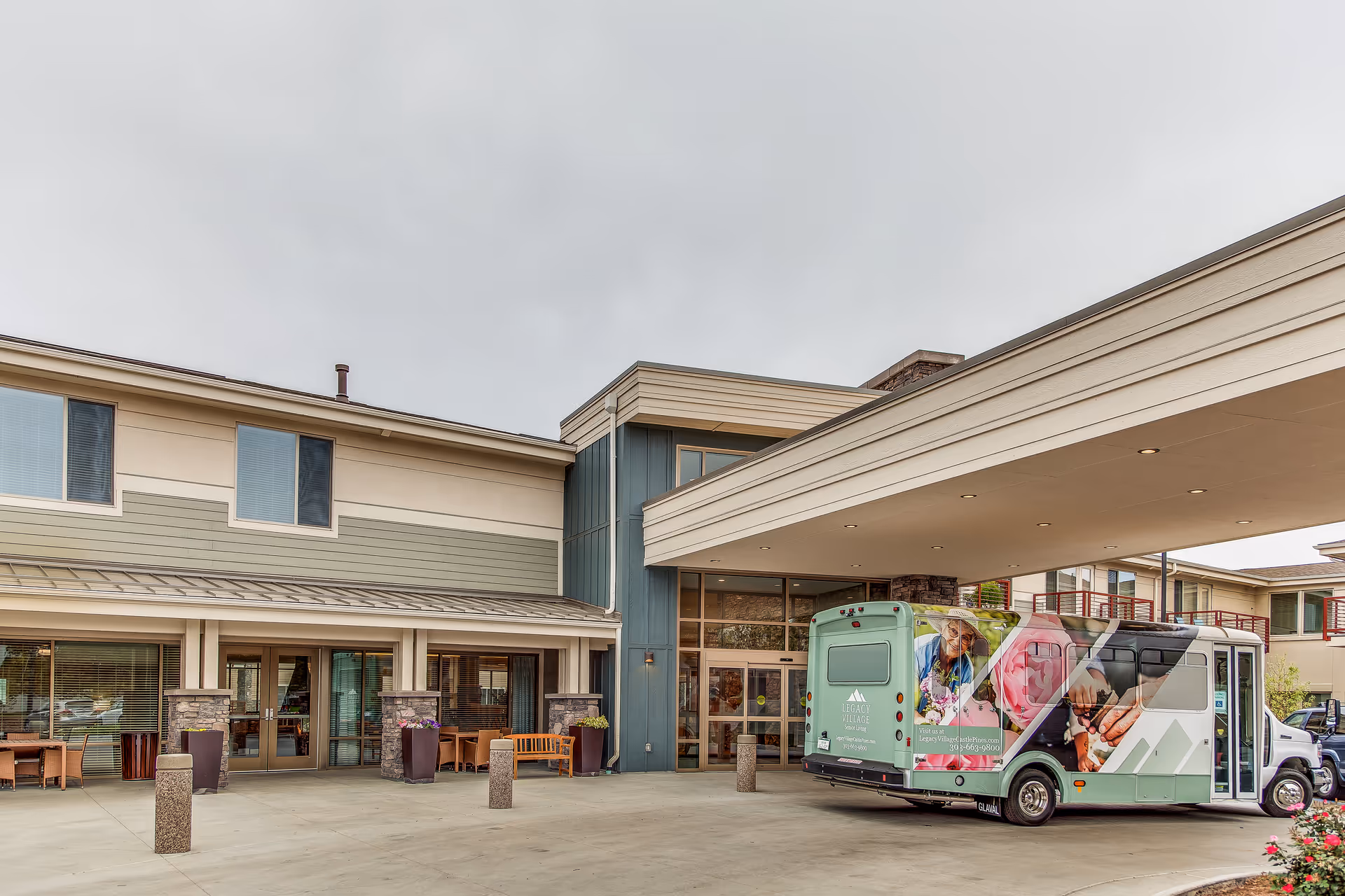 Exterior view of Legacy Village of Castle Pines showing the building entrance with a covered drop-off area. A shuttle bus with colorful images of seniors is parked under the canopy. There are benches and large planters near the entrance.