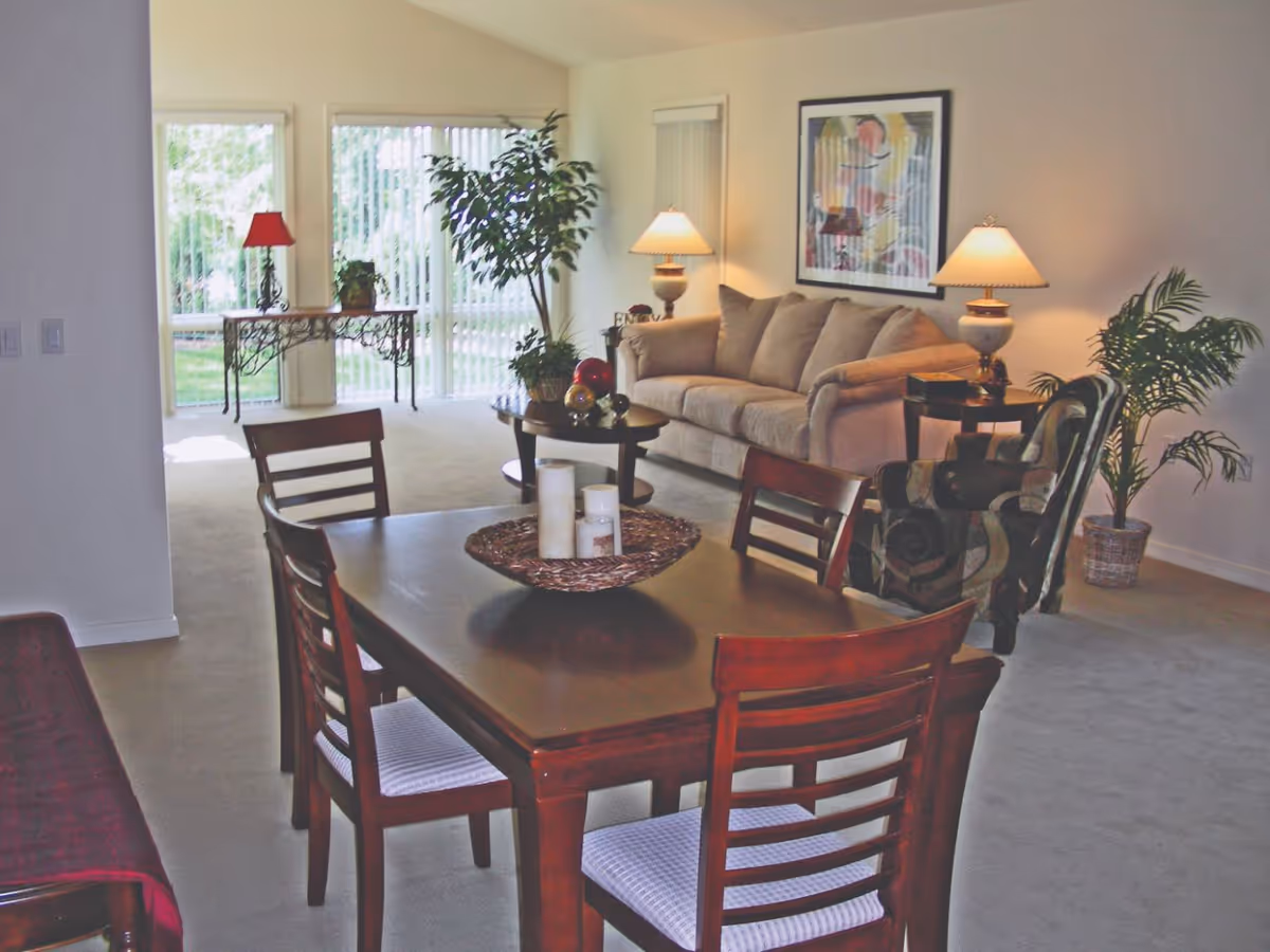 Open-plan living and dining area with a wooden dining table in the foreground, a sofa and armchair with lamps, and potted plants by sliding glass doors.