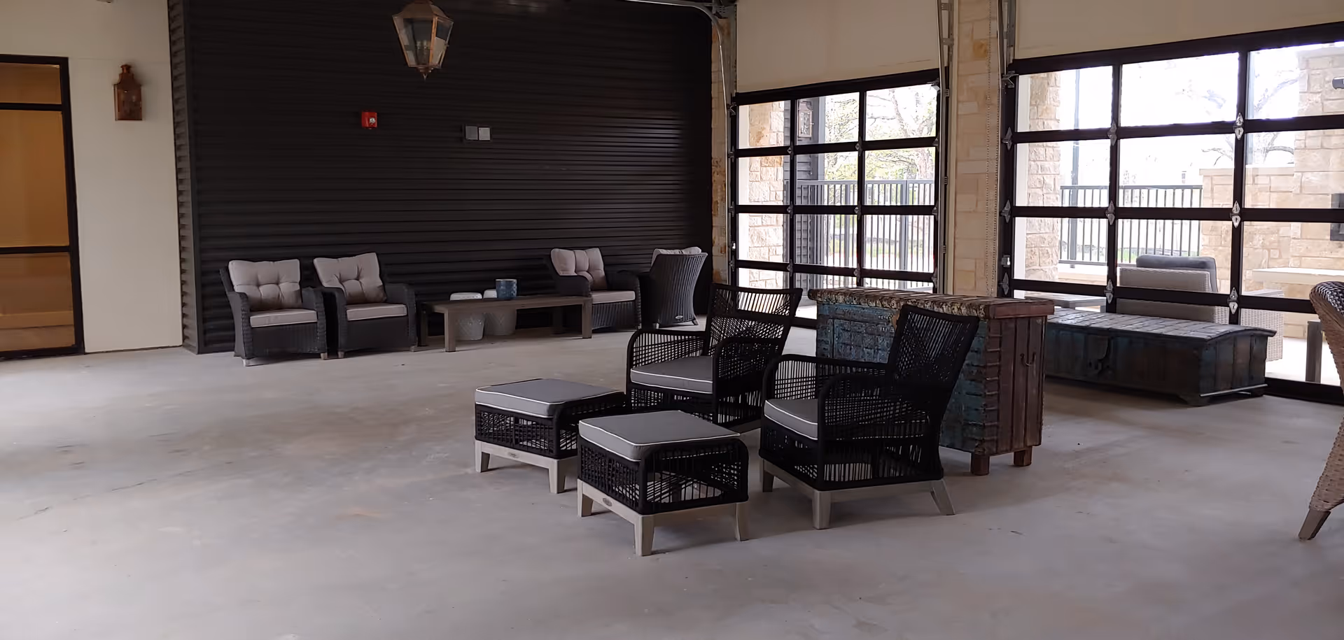 Indoor seating area with multiple cushioned chairs and ottomans arranged on a concrete floor. The space features large glass garage-style doors that open to an outdoor area with stone walls and additional seating. The interior wall is dark with a hanging light fixture, and there is a door on the left side.