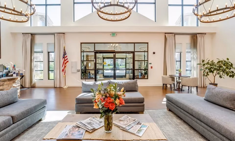 Bright and spacious senior living facility lobby with large windows, two gray sofas facing each other, a coffee table with a vase of flowers and magazines, a small round table with two chairs near the window, and an American flag near the entrance door.