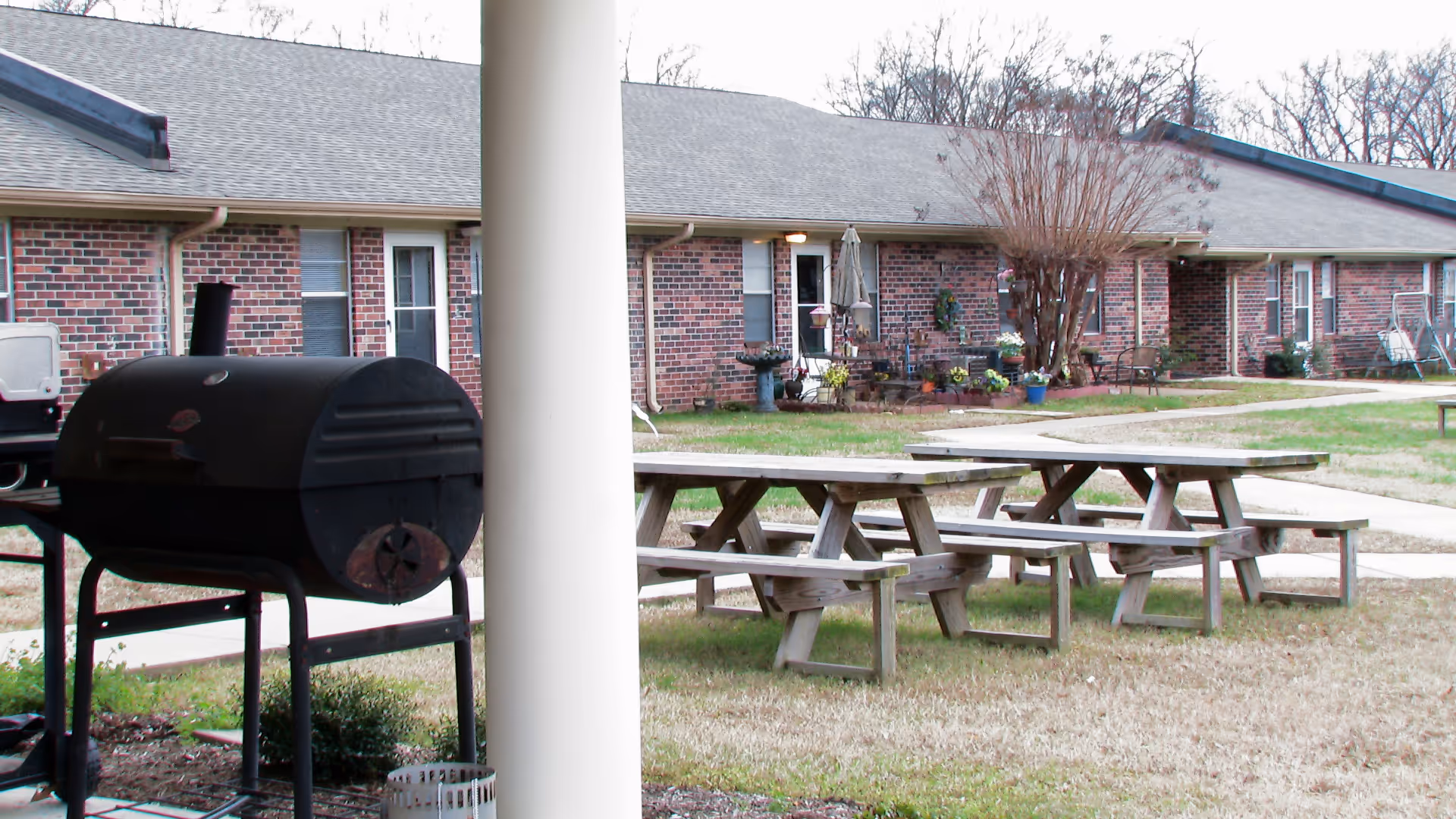 Outdoor courtyard by a single-story brick senior living building with picnic tables, a grill, and a lawn.