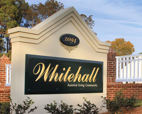 Entrance sign reading "Whitehall Assisted Living Community" with address "2094", set among brick pillars, a white fence, and landscaping.