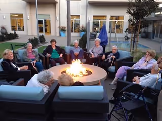 A group of elderly people sitting around a circular outdoor fire pit in a courtyard area with modern patio furniture. The setting is outside a building with large windows and doors, and there are plants and trees around the seating area.