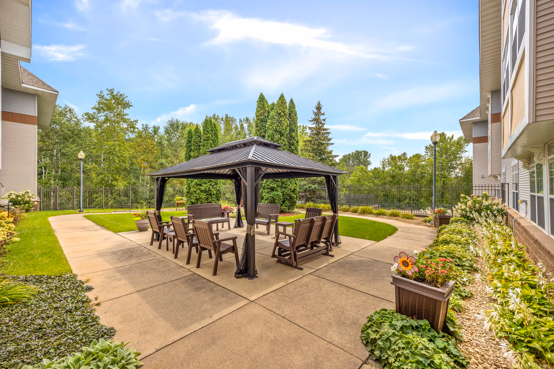 A landscaped outdoor courtyard with a central gazebo, chairs, pathways, planters and surrounding greenery between residential buildings.