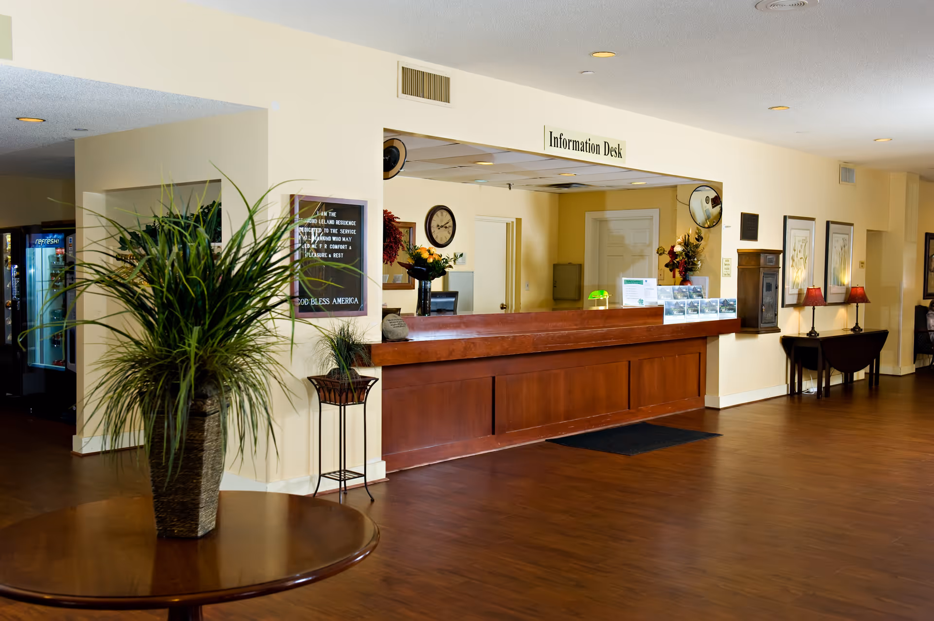 Interior view of a senior community's information desk area with a wooden reception counter, a large potted plant on a round wooden table in the foreground, framed pictures and lamps on the wall, and a vending machine in the background.