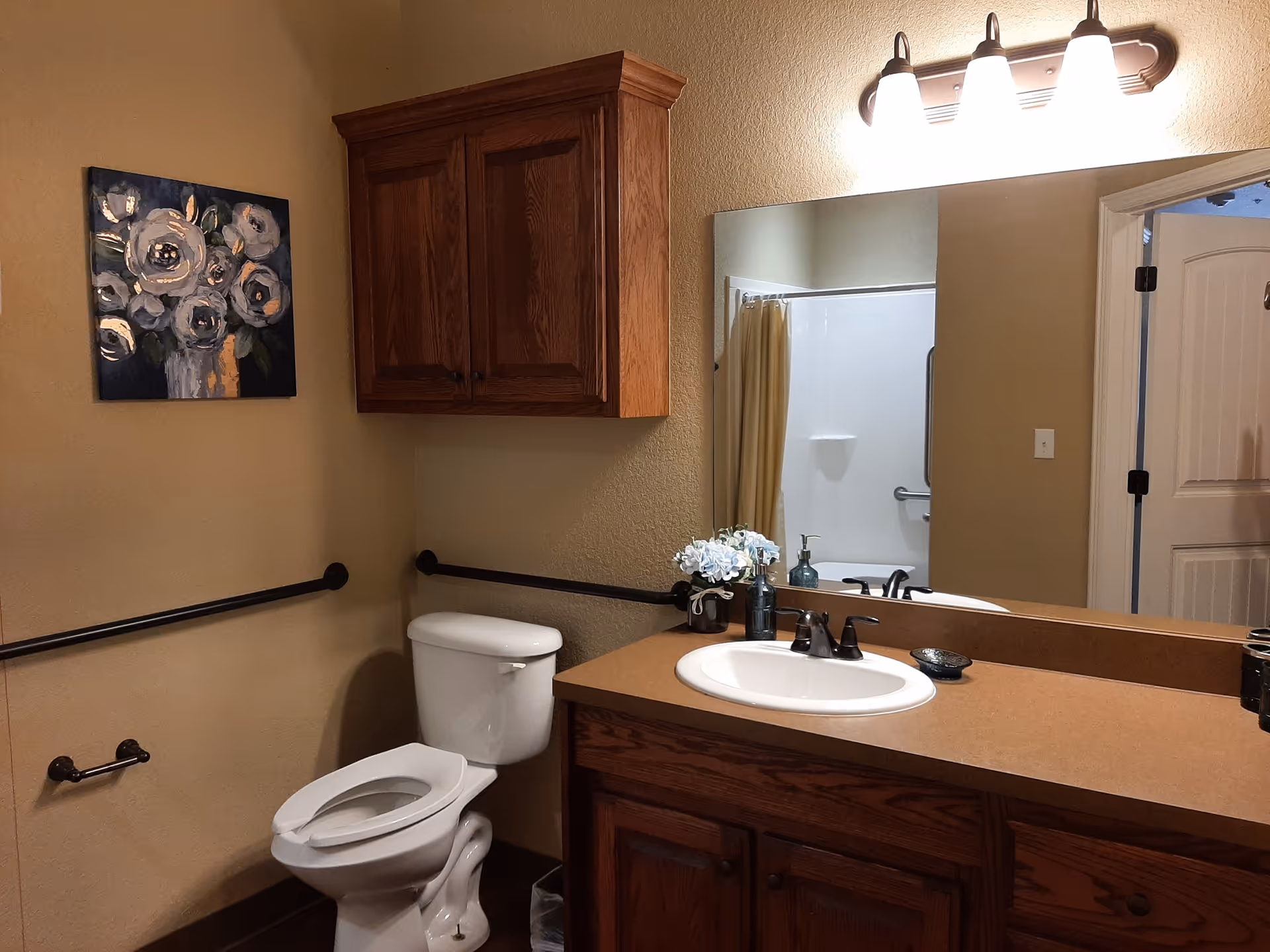 A bathroom with a white toilet, a wooden cabinet mounted on the wall, a countertop with a sink, a large mirror above the sink, and a light fixture with three bulbs. There is a painting of flowers on the wall and a shower with a yellow curtain visible in the mirror reflection.