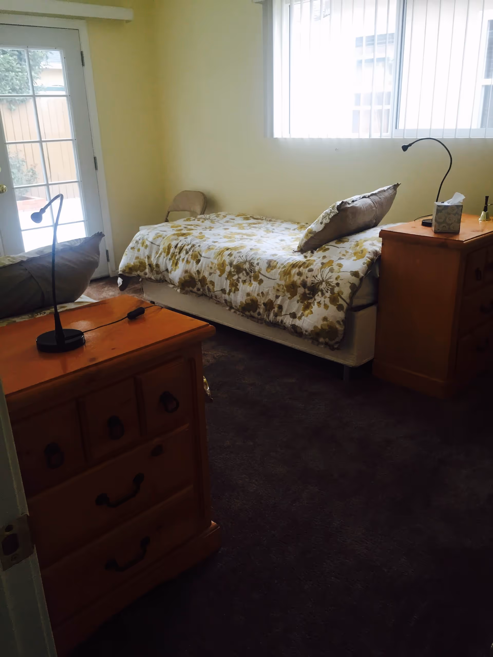 A small bedroom with a single bed covered in a floral patterned comforter and a pillow. There are two wooden dressers, one in the foreground with a black lamp on it and another beside the bed with a tissue box and a small lamp. A window with vertical blinds lets in natural light, and a glass door leads outside to a fenced area.