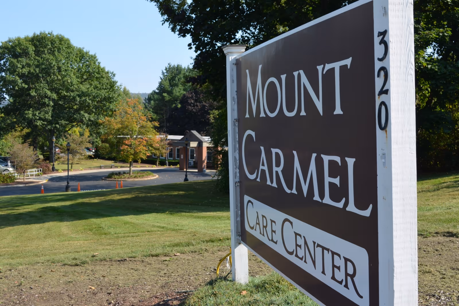 Outdoor view of the Mount Carmel Care Center sign with a driveway, trees, and a building in the background under a clear blue sky.