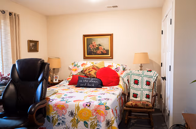 Cozy bedroom with a floral quilted bed topped with multiple pillows, a leather armchair, a wooden rocking chair draped with a crocheted blanket, bedside lamps, and a framed painting on the wall.